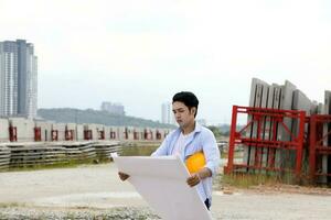 asiático hombre malayo trabajador ingeniero administración difícil la seguridad sombrero casco a construcción sitio leyendo viendo plan azul cielo foto