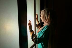 joven asiático malayo musulmán mujer vistiendo Pañuelo baju Kurung vestir a hogar en frente ventana estado animico ligero en cara oscuro antecedentes pacífico contenido calma tranquilo descanso canal preguntarse foto