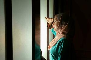 young Asian Malay muslim woman wearing headscarf baju kurung dress at home in front window mood light on face dark background peaceful content calm tranquil break trough wonder photo