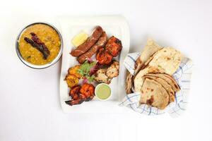 Mixed grill kebab chicken mutton beef prawn fish mint sauce lemon pickled onion bread basket chapati naan tandoori paratha dal lentil fry tadka copper pot on white dish over white background photo