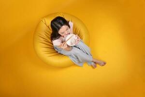 Beautiful young south east Asian woman holding empty chopstick soup spoon bowl posing see eat taste smell feed offer satisfaction yummy siting on beanbag seat yellow orange background top view photo