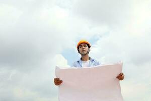 asiático hombre malayo trabajador ingeniero administración difícil la seguridad sombrero casco a construcción sitio leyendo viendo plan azul cielo foto