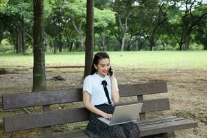 Young asian chinese woman outdoor on park bench use laptop computer think read look talk on phone photo