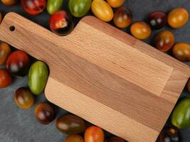 Red orange yellow green tomato mix variety on around wooden chopping board frame copy text space over black slate stone background photo