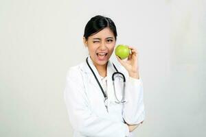 Young Asian female doctor wearing apron uniform tunic stethoscope holding pointing showing eating healthy green apple photo