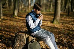 Autumn outdoor portrait of teenager boy sit in stump and drink tea from thermos cup in forest. photo