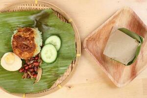 Nasi Lemak fragrant rice cooked in coconut milk served with sambal friend peanut anchovy boiled egg packed in banana leaf on round bamboo plate on wooden background photo