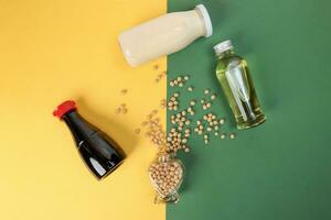 Soyabeans pouring splashing from hart shape glass bottle around bottle of soya bean milk oil sauce on yellow green background photo