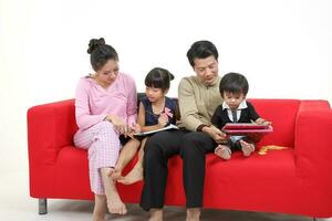 South East Asian young couple father mother son daughter parent girl boy child on red sofa read write study book use computer talk phone play tab on white background photo