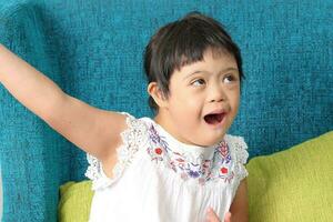 Sureste asiático pequeño pequeño niña niño sentado en azul sofá Mira contento sonrisa actitud abierto brazo contento abrazo cantar. ella tener abajo síndrome foto