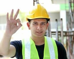 South East Asian young Malay man wearing yellow safety helmet vest looking at camera ok finger sign photo
