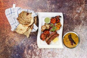 Mixed grill kebab chicken mutton beef prawn fish mint sauce lemon pickled onion bread basket chapati naan tandoori paratha dal lentil fry tadka copper pot on white dish over rustic background photo