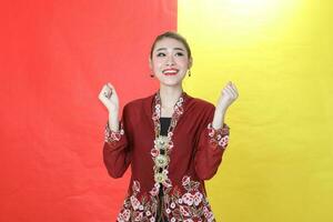 Asian woman traditional maroon kebaya sharong on red yellow paper background posing happy success fist in air photo