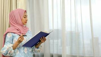 young Asian Malay Muslim woman wearing headscarf at home office next to window look at book document study think write photo