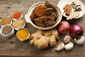 Beef bhuna dry curry with potato in ceramic bowl ingredients chilli cumin coriander turmeric powered salt onion garlic ginger bay leaf cinnamon cardamom clove over rustic wood background photo
