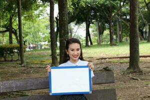 Young asian chinese woman outdoor on park bench hold whiteboard copy text space smile look point photo