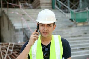 South East Asian young Malay man wearing white safety helmet yellow vest talking on phone looking front at camera photo