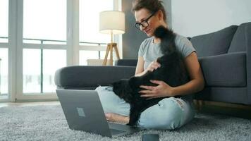 mine de rien habillé femme avec des lunettes est assis sur une tapis avec une ordinateur portable, détient sur sa les genoux et coups une duveteux chat et travaux dans une confortable chambre. éloigné travail à l'extérieur le Bureau video