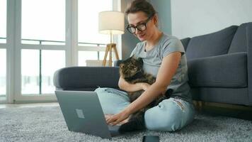 Casually dressed woman with glasses sits on a carpet with a laptop, holds on her knees and strokes a fluffy cat and works in a cozy room. Remote work outside the office video