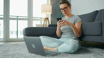 Casually dressed woman sitting on carpet with laptop and smartphone and working in cozy room. Remote work outside the office video