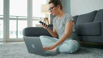 mine de rien habillé femme séance sur tapis avec portable et téléphone intelligent et travail dans confortable chambre. éloigné travail à l'extérieur le Bureau video