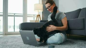 Casually dressed woman with glasses sits on a carpet with a laptop, holds on her knees and strokes a fluffy cat and works in a cozy room. Remote work outside the office video