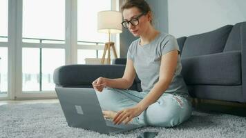 Casually dressed woman with glasses is sitting on carpet with laptop in cozy room. She communicates and works remotely. Remote work outside the office video