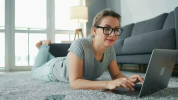 donna con bicchieri è dire bugie su il pavimento e Lavorando su un' il computer portatile. concetto di a distanza opera video