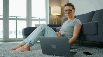 por casualidad vestido mujer con lentes es sentado en alfombra con ordenador portátil en acogedor habitación. ella comunica y trabajos remotamente remoto trabajo fuera de el oficina video