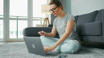 Woman with glasses is sitting on the floor and makes an online purchase using a credit card and laptop video