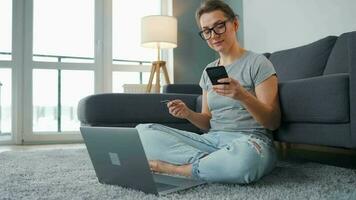 mujer con lentes es sentado en el alfombra y hace un en línea compra utilizando un crédito tarjeta y teléfono inteligente en línea compras, estilo de vida tecnología video