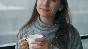 caucásico mujer corsé en balcón durante nevada con taza de caliente café o té. ella mira a el copos de nieve y respira video