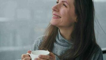 Kaukasisch vrouw blijft Aan balkon gedurende sneeuwval met kop van heet koffie of thee. ze looks Bij de sneeuwvlokken en ademt video