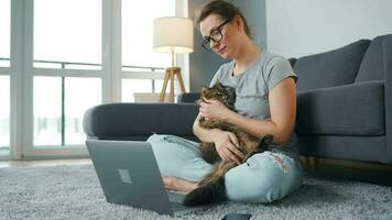 por casualidad vestido mujer con lentes se sienta en un alfombra con un computadora portátil, sostiene en su rodillas y golpes un mullido gato y trabajos en un acogedor habitación. remoto trabajo fuera de el oficina video