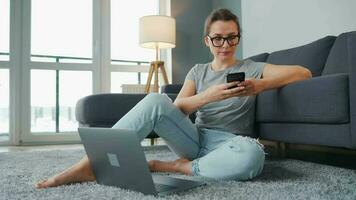 mine de rien habillé femme séance sur tapis avec portable et téléphone intelligent et travail dans confortable chambre. éloigné travail à l'extérieur le Bureau video