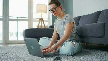 por casualidad vestido mujer con lentes es sentado en alfombra con ordenador portátil en acogedor habitación. ella comunica y trabajos remotamente remoto trabajo fuera de el oficina video