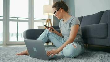 mine de rien habillé femme séance sur tapis avec portable et téléphone intelligent et travail dans confortable chambre. éloigné travail à l'extérieur le Bureau video