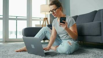 Casually dressed woman sitting on carpet with laptop and smartphone and working in cozy room. Remote work outside the office video