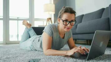 donna con bicchieri è dire bugie su il pavimento e Lavorando su un' il computer portatile. concetto di a distanza opera video