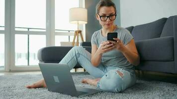 mine de rien habillé femme séance sur tapis avec portable et téléphone intelligent et travail dans confortable chambre. éloigné travail à l'extérieur le Bureau video