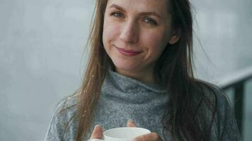 Kaukasisch vrouw blijft Aan balkon gedurende sneeuwval met kop van heet koffie of thee. ze looks Bij de sneeuwvlokken en ademt video