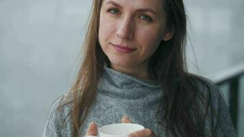 Kaukasisch vrouw blijft Aan balkon gedurende sneeuwval met kop van heet koffie of thee. ze looks Bij de sneeuwvlokken en ademt video