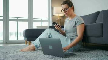 Casually dressed woman sitting on carpet with laptop and smartphone and working in cozy room. Remote work outside the office video