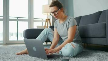 beiläufig gekleidet Frau Sitzung auf Teppich mit Laptop und Smartphone und Arbeiten im gemütlich Zimmer. Fernbedienung Arbeit draußen das Büro video