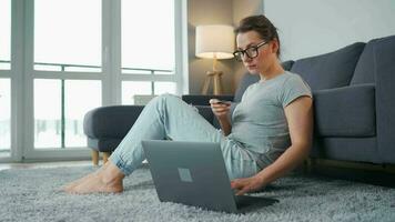 mujer con lentes es sentado en el piso y hace un en línea compra utilizando un crédito tarjeta y ordenador portátil video