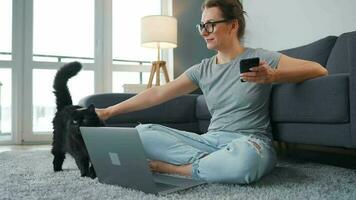 Casually dressed woman with glasses sits on a carpet with a laptop, holds on her knees and strokes a fluffy cat and works in a cozy room. Remote work outside the office video
