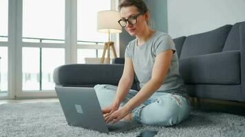 beiläufig gekleidet Frau Sitzung auf Teppich mit Laptop und Smartphone und Arbeiten im gemütlich Zimmer. Fernbedienung Arbeit draußen das Büro video