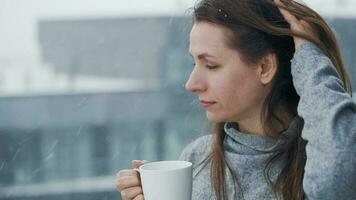 caucasian kvinna vistelser på balkong under snöfall med kopp av varm kaffe eller te. hon utseende på de snöflingor och andas video