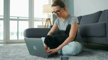 mine de rien habillé femme avec des lunettes est assis sur une tapis avec une ordinateur portable, détient sur sa les genoux et coups une duveteux chat et travaux dans une confortable chambre. éloigné travail à l'extérieur le Bureau video
