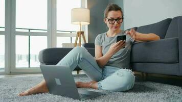 mine de rien habillé femme séance sur tapis avec portable et téléphone intelligent et travail dans confortable chambre. éloigné travail à l'extérieur le Bureau video